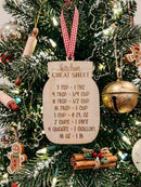 A wooden kitchen cheat sheet Christmas ornament hanging on a decorated Christmas tree. The ornament is shaped like a mason jar and features engraved conversion charts for cups, pints, quarts, and pounds, with a red and white gingham ribbon hanging from it.