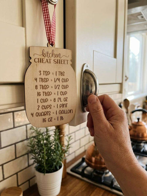 A hand holding a round metal keyring with a wooden kitchen cheat sheet ornament hanging on a cabinet in a real kitchen setting.