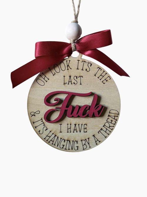 A round wooden hanging ornament with engraved black text reading 'OH LOOK IT'S THE LAST FUCK & IT'S HANGING BY A THREAD' and a raised dark red 'FUCK' in the center, tied with a dark red ribbon.